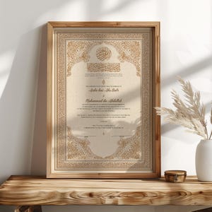 May include: Framed marriage certificate with ornate gold detailing and Arabic calligraphy. The document features the names Aisha bint Abu Bakr and Muhammad ibn Abdullah. The frame is a light brown color and sits on a wooden surface.