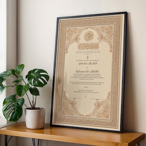May include: Framed certificate with ornate gold and beige detailing, featuring Arabic calligraphy and English text. The black frame surrounds the document, which is leaning against a wall. A potted plant sits on a wooden surface in the foreground.