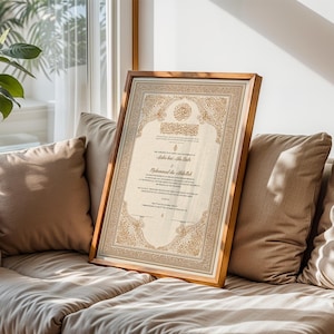 May include: A framed, ornate document with Arabic calligraphy and decorative borders. The frame is a warm brown, and the document features gold and beige tones. The artwork is leaning against a beige sofa, near a window with natural light.