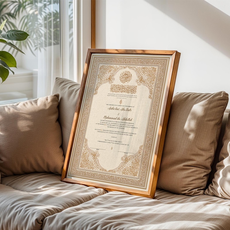May include: A framed, ornate document with Arabic calligraphy and decorative borders. The frame is a warm brown, and the document features gold and beige tones. The artwork is leaning against a beige sofa, near a window with natural light.