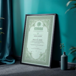 May include: Framed Islamic marriage certificate with ornate green border and text. The certificate is in a black frame, leaning against a teal wall. A small vase with greenery sits nearby. The certificate features Arabic calligraphy and English text.