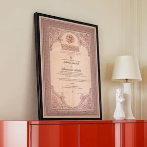 May include: Framed document with ornate red and gold detailing. The document features Arabic calligraphy and text, set within a black frame. The framed artwork is displayed on a red cabinet, with a white lamp and decorative figurine in the background.