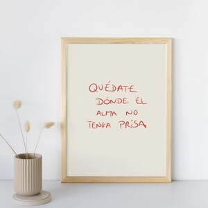 Puede incluir: Un cuadro enmarcado con el texto manuscrito en rojo "QUÉDATE DONDE EL ALMA NO TENGA PRISA". El marco es de madera clara y el fondo es blanco roto. A la izquierda, un jarrón de cerámica beige con flores secas.