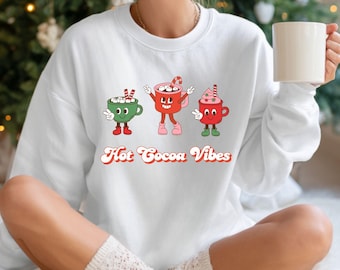 May include: White sweatshirt with a graphic of three cartoon cocoa mugs in green, red, and pink, each with a different design. The text "Hot Cocoa Vibes" is printed below the mugs in a retro font.