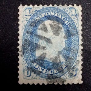 Puede incluir: Un sello postal vintage azul y blanco. El sello presenta un retrato ovalado de una persona, con las palabras "POSTAGE" y "ONE CENT" alrededor del retrato. El sello tiene un borde detallado y estampado.