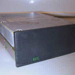 Könnte beinhalten: Rechteckförmige, silberfarbene Festplatte mit schwarzer Frontplatte. Die Festplatte ist in einem Metallrahmen montiert und hat einen kleinen grünen Aufkleber. Es handelt sich um ein älteres Modell.