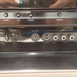 May include: A black audio receiver front panel featuring buttons labeled TAPE, TUNER, CD, PHONO, and DISPLAY. The interface includes a navigation pad, a SETUP MIC port, an S-VIDEO port, a VIDEO port, L-AUDIO-R ports, and a digital input port.