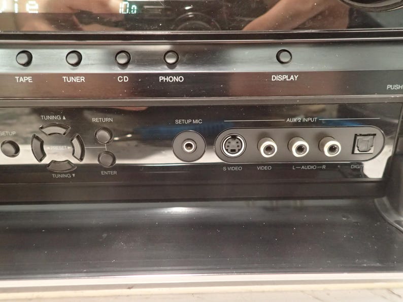 May include: A black audio receiver front panel featuring buttons labeled TAPE, TUNER, CD, PHONO, and DISPLAY. The interface includes a navigation pad, a SETUP MIC port, an S-VIDEO port, a VIDEO port, L-AUDIO-R ports, and a digital input port.