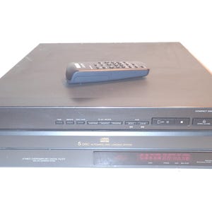 Puede incluir: Un reproductor de discos compactos Sony CDP-C300M negro con un mando a distancia en la parte superior. El reproductor tiene un sistema de carga automática de 5 discos y una pantalla digital. El mando es negro con varios botones.