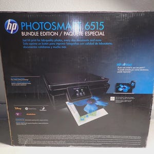 Peut inclure: Une boîte noire d'imprimante HP Photosmart 6515 édition groupée. La boîte présente le logo HP et le texte "PHOTOSMART 6515 BUNDLE EDITION / PAQUETE ESPECIAL". La boîte comprend également le texte "Just hit print for lab-quality photos, every day documents and more".