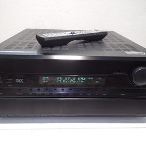 May include: A black Onkyo audio-video receiver with a digital display showing FM 87.5 MHz and PLIIx Movie. The front panel features a large volume knob, multiple input buttons, and a THX logo. A black remote control rests on the top vented surface.