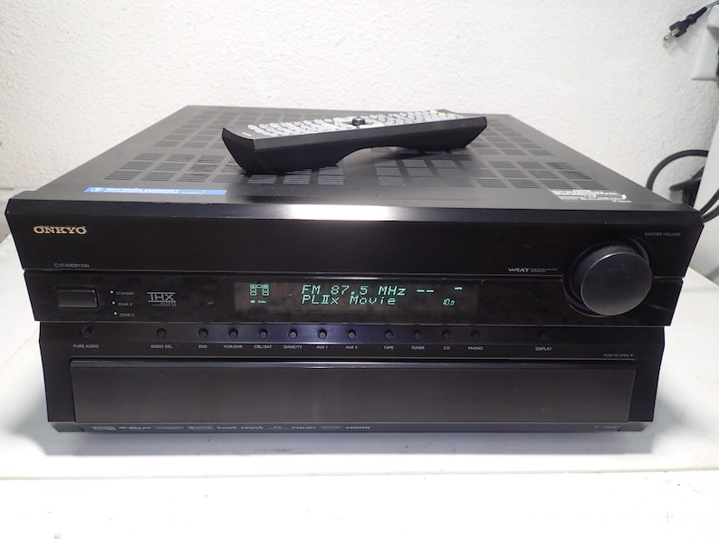May include: A black Onkyo audio-video receiver with a digital display showing FM 87.5 MHz and PLIIx Movie. The front panel features a large volume knob, multiple input buttons, and a THX logo. A black remote control rests on the top vented surface.