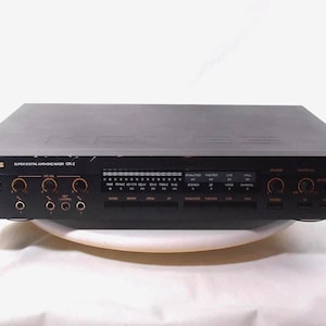 Puede incluir: Mezclador de karaoke digital Konės Super Digital CR-2 negro. El panel frontal tiene varias perillas y botones con detalles en naranja. El mezclador tiene un diseño elegante y rectangular y probablemente se usa para mezclar audio y karaoke.