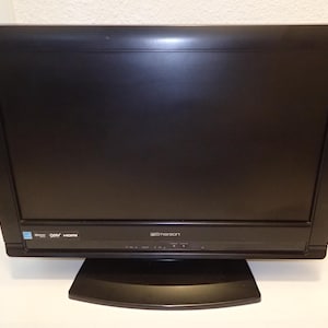 May include: A black Emerson television with a wide rectangular screen and a matching black base stand. The front panel features the Emerson brand logo, along with small printed labels for HDMI and other connectivity ports located at the bottom.