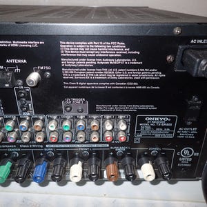 May include: The back panel of a black Onkyo AV receiver model TX-SR805 featuring numerous color-coded RCA input and output ports, speaker wire terminals, an antenna connection, and an AC power inlet with a connected black power cord.