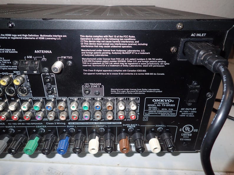 May include: The back panel of a black Onkyo AV receiver model TX-SR805 featuring numerous color-coded RCA input and output ports, speaker wire terminals, an antenna connection, and an AC power inlet with a connected black power cord.