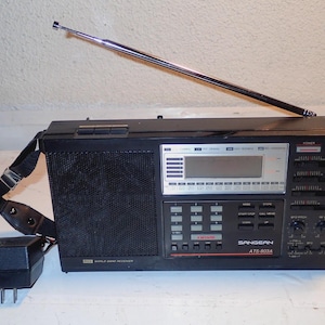 Op de afbeelding: Zwarte Sangean ATS-803A wereldontvanger met een zilveren antenne en een zwarte schouderriem. De radio heeft een digitaal display en diverse knoppen en draaiknoppen. Een AC-adapter is aangesloten.