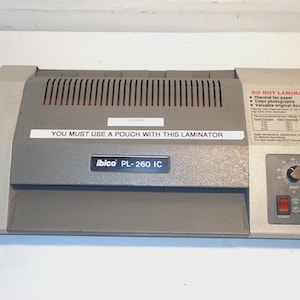 Puede incluir: Una plastificadora Ibico PL-260 IC gris y beige. La máquina tiene un panel de control con un dial y dos botones rojos. El texto en la máquina incluye "YOU MUST USE A POUCH WITH THIS LAMINATOR" y "DO NOT LAMINATE".