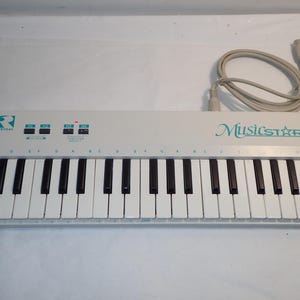 May include: A white MusicStar keyboard with black and white keys. The keyboard has a series of buttons and a power indicator light. The text "MusicStar" is in teal script. A gray cable is connected to the keyboard.