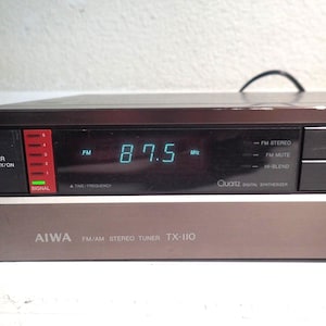 以下が含まれることがあります： 黒とシルバーのAIWA FM/AMステレオチューナーTX-110。デジタルディスプレイには青色で「87.5 MHz」と表示されています。チューナーには電源ボタン、信号インジケーター、プリセットボタンがあります。「Quartz Digital Synthesizer」の文字も表示されています。