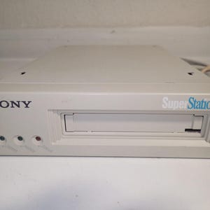 Op de afbeelding: Een vintage witte Sony Super Station videogameconsole. De console heeft een sleuf voor het plaatsen van een spelcartridge en heeft het Sony-logo links en "Super Station" in blauwe tekst rechts. Het heeft drie indicatielampjes en een netsnoer.