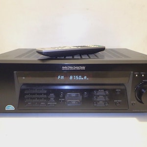 Pode incluir: Receptor Sony Audio/Video Control Center preto com um controle remoto em cima. O receptor exibe "FM 8750.A" no painel frontal. Possui vários botões, um botão de volume e conectores de entrada.