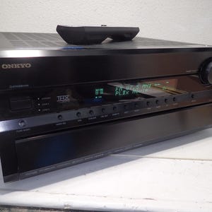 May include: A black Onkyo audio-video receiver with a THX logo, a digital display showing FM 87.5 MHz and PLIIx Movie, a large volume knob on the right, and a row of control buttons. A black remote control rests on the top surface of the unit.