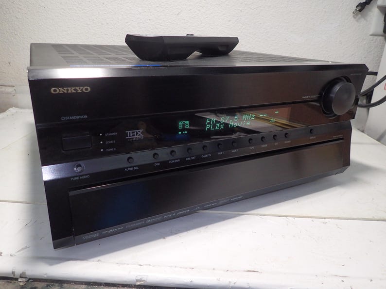 May include: A black Onkyo audio-video receiver with a THX logo, a digital display showing FM 87.5 MHz and PLIIx Movie, a large volume knob on the right, and a row of control buttons. A black remote control rests on the top surface of the unit.