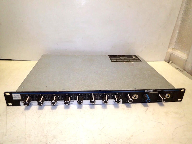 May include: A silver and black Shure SC3400 audio mixer with multiple knobs and input jacks. The mixer has a black front panel with blue accents and white text. The device is designed for professional audio applications.