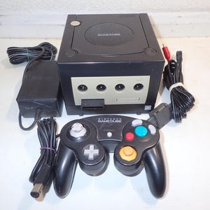 Può includere: Una console per videogiochi Nintendo GameCube nera con controller nero abbinato, alimentatore e cavo AV. La console presenta un pannello frontale con quattro porte per controller e uno slot per schede di memoria, con il logo GameCube.