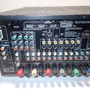 May include: The back panel of a black audio-video receiver featuring numerous input and output ports, including HDMI, component video, coaxial, optical, and speaker terminals. The panel is labeled with various connection types and technical text.