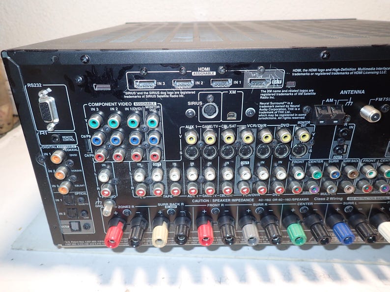 May include: The back panel of a black audio-video receiver featuring numerous input and output ports, including HDMI, component video, coaxial, optical, and speaker terminals. The panel is labeled with various connection types and technical text.