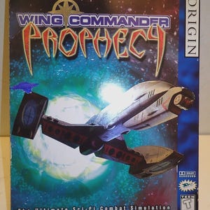 May include: A Wing Commander Prophecy video game box. The box features a spaceship design with a blue and white background. The game title is in gold and red lettering. The text "The Ultimate Sci-Fi Combat Simulation" is also visible.