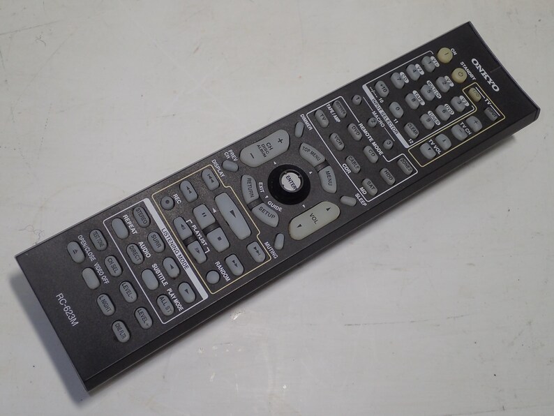 May include: A black Onkyo RC-623M remote control featuring a rectangular body with numerous grey buttons for device operation, including volume, playback, and input selection controls, with white text labels printed on the surface of the device.