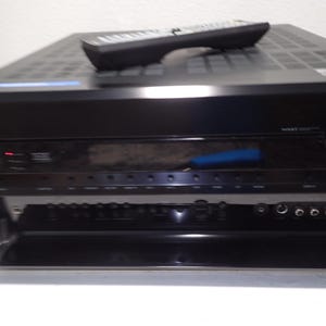 May include: A black Onkyo audio-video receiver with a remote control resting on top. The front panel features a large volume knob, a digital display, and a fold-down door revealing additional input ports, buttons, and a headphone jack.