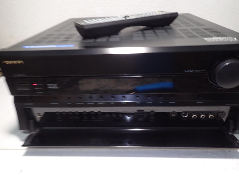 May include: A black Onkyo audio-video receiver with a remote control resting on top. The front panel features a large volume knob, a digital display, and a fold-down door revealing additional input ports, buttons, and a headphone jack.