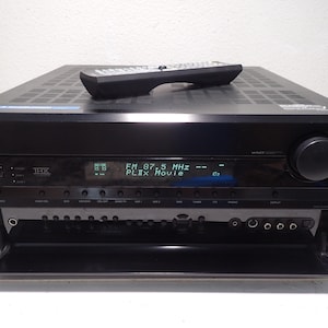 May include: A black Onkyo audio-video receiver with a digital display showing FM 87.5 MHz and PLIIx Movie. The unit features a large volume knob, a THX logo, and a drop-down front panel revealing various input ports. A black remote control rests on top.