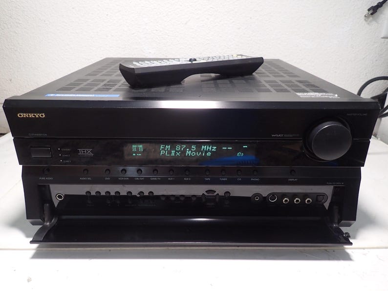 May include: A black Onkyo audio-video receiver with a digital display showing FM 87.5 MHz and PLIIx Movie. The unit features a large volume knob, a THX logo, and a drop-down front panel revealing various input ports. A black remote control rests on top.