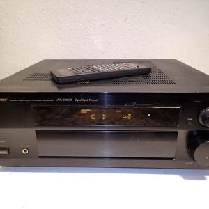 Pode incluir: Receptor de processador de sinal digital Pioneer VSX-D810S preto com um controle remoto em cima. O receptor tem um display mostrando "CD" e "-49". O painel frontal tem um botão de energia, uma tomada para fone de ouvido e um controle de volume.