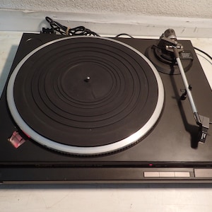 Puede incluir: Tocadiscos negro vintage con un plato con borde plateado y un brazo fonocaptor. El tocadiscos tiene un cuerpo negro con botones plateados y una luz indicadora de encendido roja. Dispositivo de audio clásico.