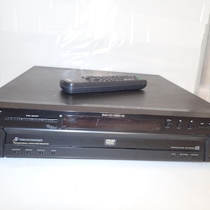 Puede incluir: Un cambiador de DVD/CD de 5 discos Sony en color negro con un mando a distancia a juego encima. El panel frontal incluye un botón de encendido, controles de selección de disco, una pantalla central, botones de reproducción y el texto DVD/CD/VIDEO CD y DVP-NC665.