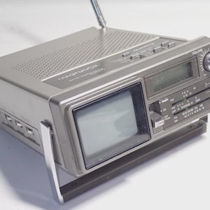 Puede incluir: Unidad combinada de TV/radio portátil Magnavox plateada. Cuenta con una pantalla pequeña, perillas de volumen y sintonización, y una antena telescópica. La unidad tiene un asa y varios botones de control.