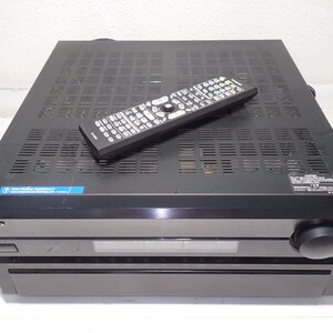 May include: A black Onkyo audio-video receiver with a rectangular chassis, a front-facing volume knob, a digital display panel, and a power button. A black remote control with numerous buttons is placed on top of the ventilated metal casing.