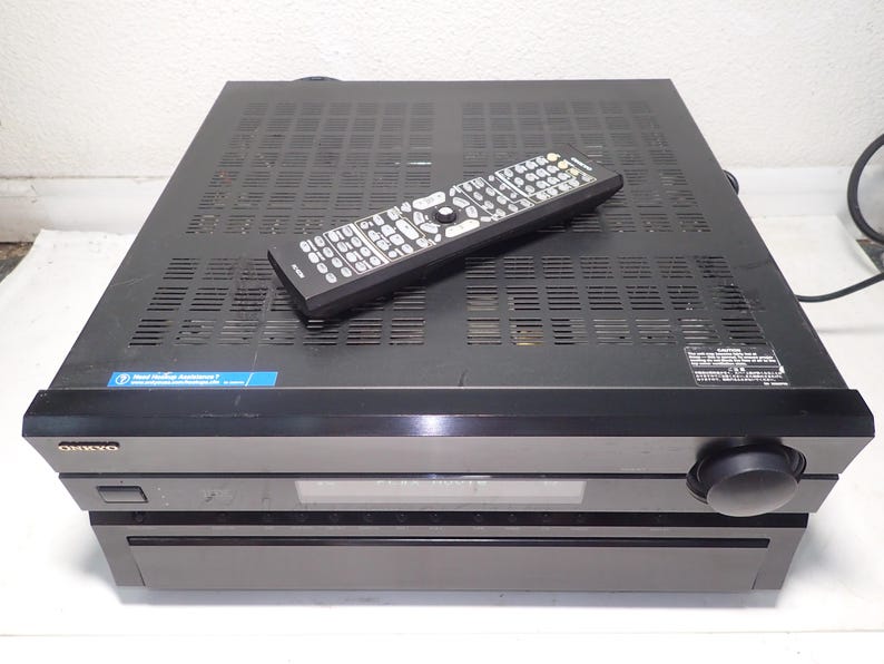 May include: A black Onkyo audio-video receiver with a rectangular chassis, a front-facing volume knob, a digital display panel, and a power button. A black remote control with numerous buttons is placed on top of the ventilated metal casing.