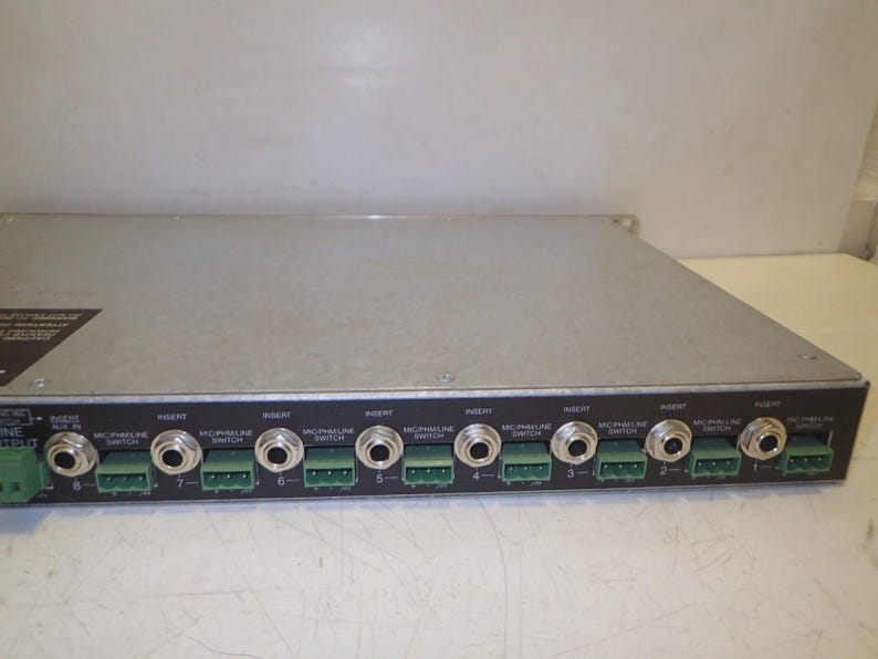 May include: A silver-colored audio mixer with multiple input and output jacks. The front panel features numerous ports labeled "Insert" and "Mic/Phaline Switch," along with green connectors. The device is designed for professional audio applications.