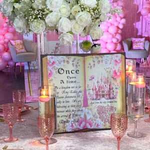 May include: A fairytale-themed table setting with a large floral arrangement of white roses. A decorative book with the text "Once Upon A Time" and a castle illustration is the centerpiece. Pink goblets, plates, and floating candles complete the elegant decor.