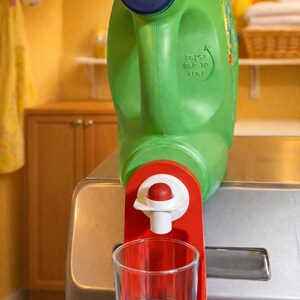 May include: A green laundry detergent bottle with a blue cap and the text "TWIST CAP TO VENT." A red dispenser with a clear glass cup is attached to the bottle. The dispenser is designed to measure and dispense the detergent.