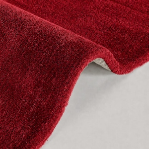 May include: A deep red rug with a thick, plush pile texture. The rug is folded over to reveal a light-colored, woven backing material. The surface of the rug features a dense, soft fiber construction with a consistent, solid crimson color throughout.
