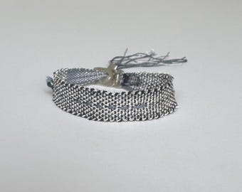 May include: A woven bracelet in shades of gray and silver, featuring a textured pattern. The bracelet has a silver-colored clasp and delicate thread tassels at the end. The bracelet is a stylish accessory.