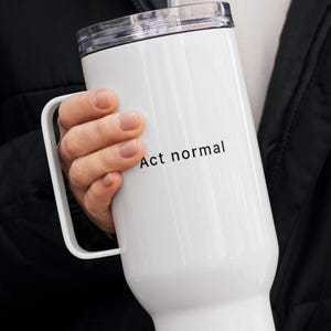 Peut inclure: Gobelet blanc avec une poignée et un couvercle transparent. Le gobelet porte l'inscription "Act normal" en noir. Une paille transparente est insérée dans le couvercle.